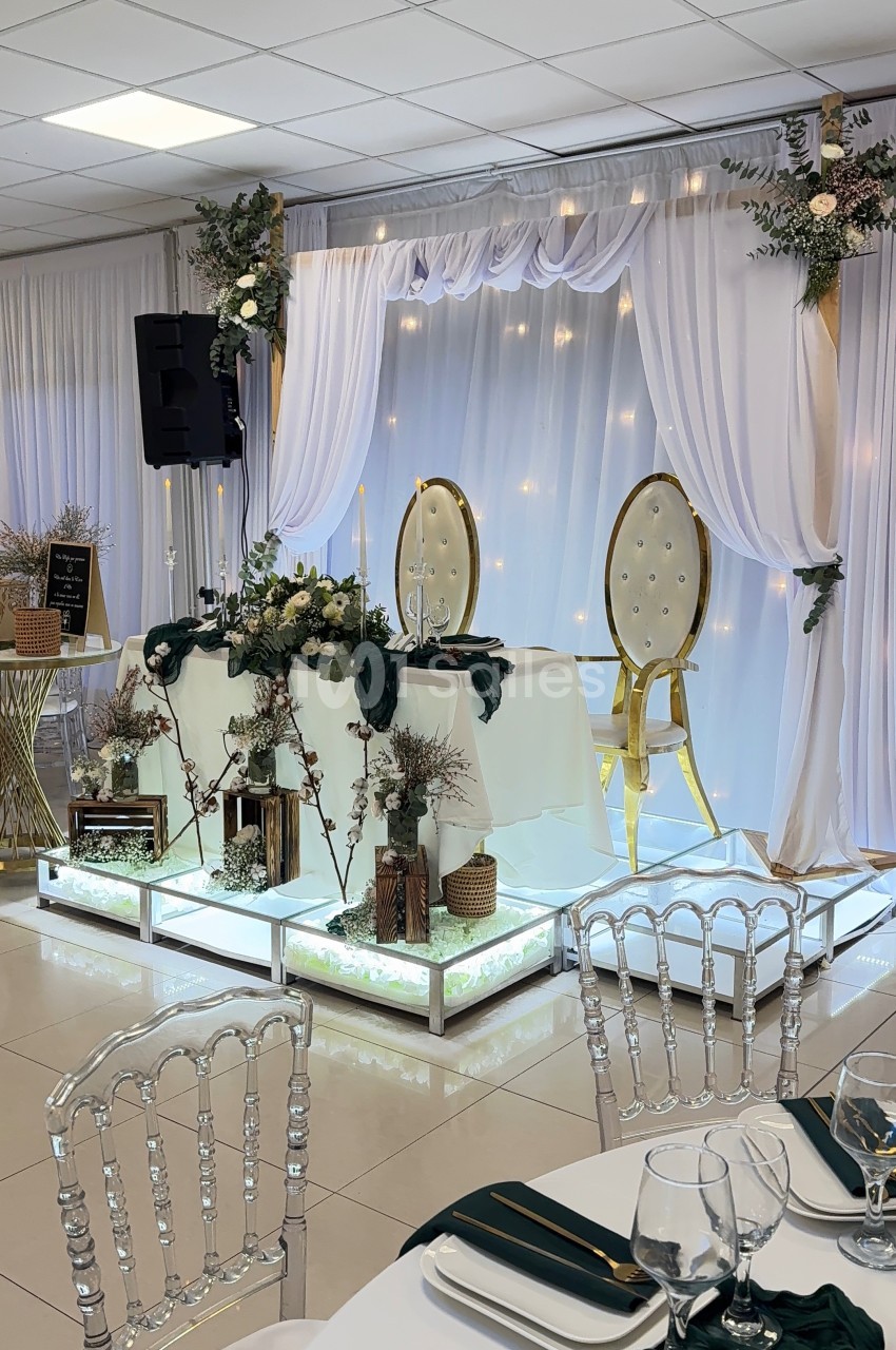 Décoration de table de mariage avec chaises dorées, rideaux blancs, guirlandes lumineuses et détails floraux.