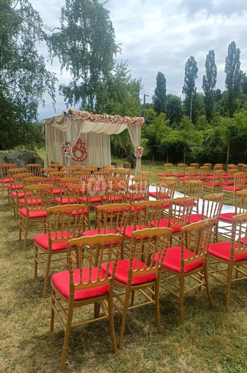 Chaises rouges alignées en extérieur devant une arche décorée pour une cérémonie, entourées de verdure.