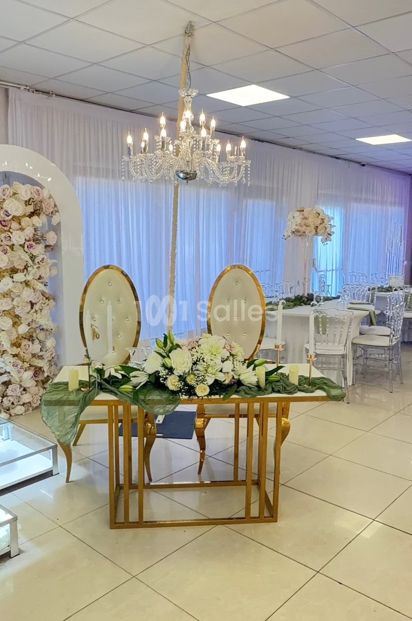 Table décorée pour un mariage avec fleurs blanches, chandeliers et fauteuils, dans une salle lumineuse ornée de rideaux…