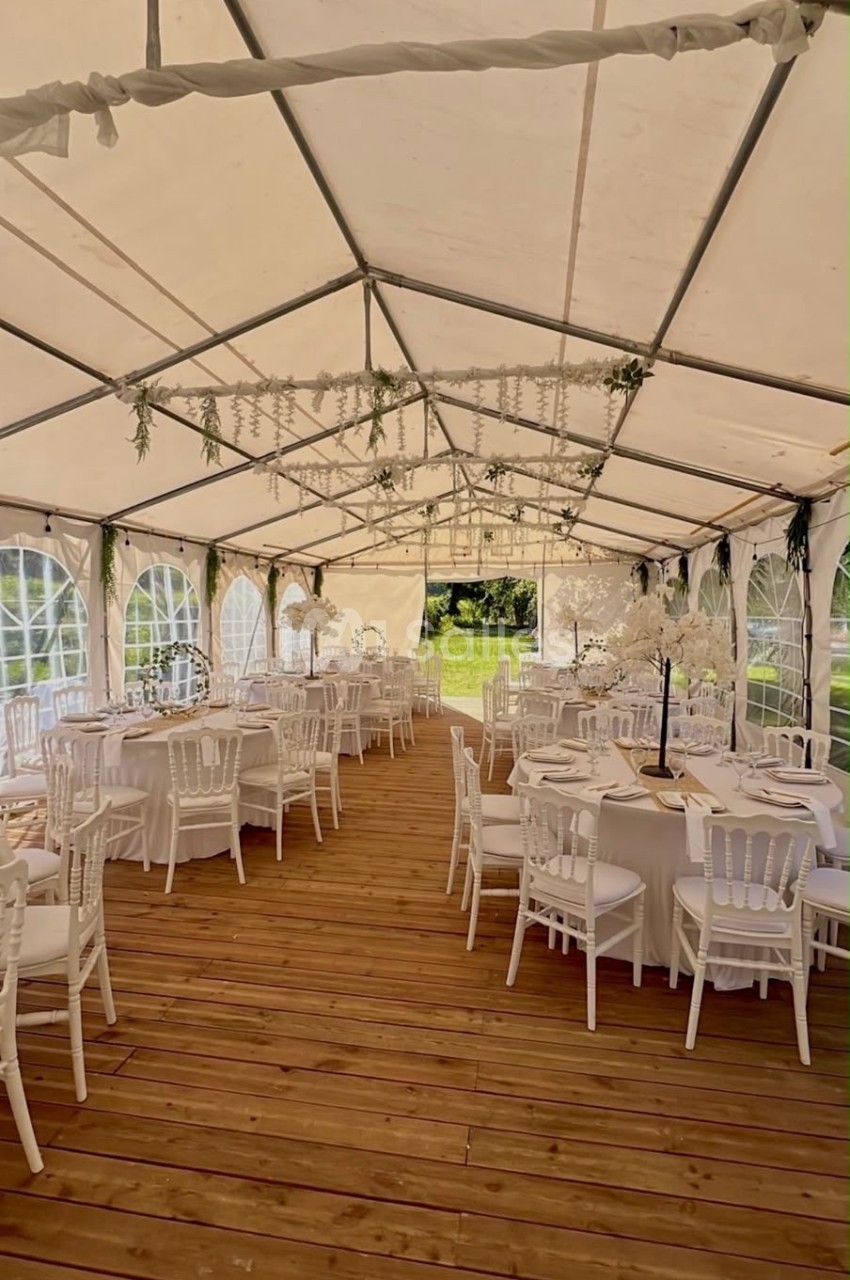 Intérieur d'une tente de réception avec tables rondes dressées, chaises blanches et décorations florales suspendues.