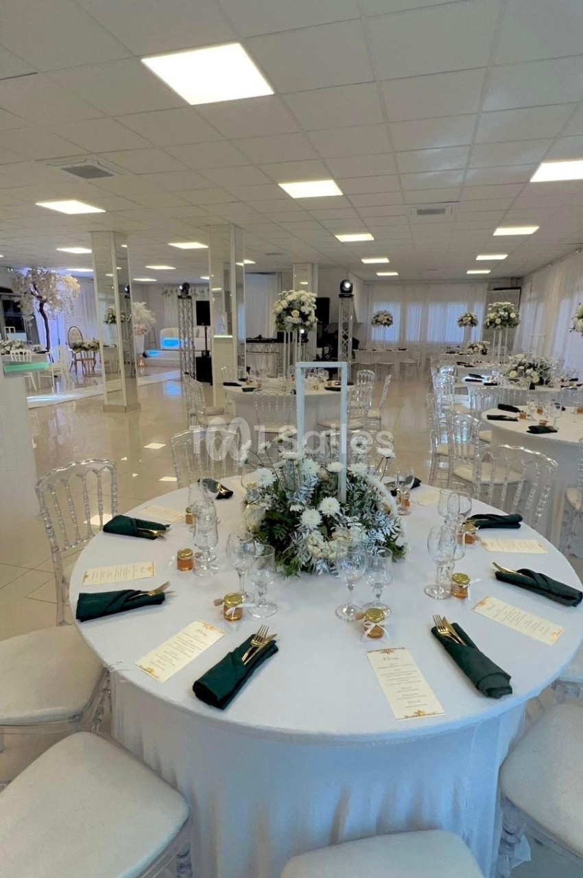 Salle de réception lumineuse avec tables rondes décorées de centres de table floraux, nappes blanches et serviettes vertes.