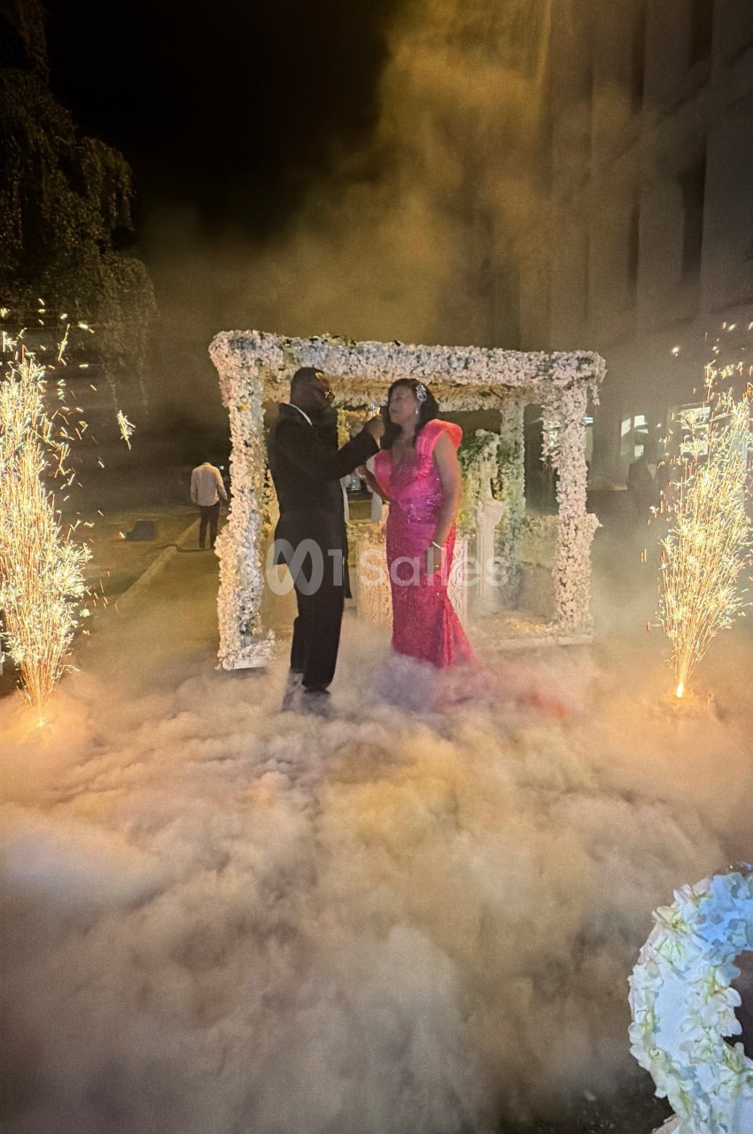 Un couple danse sous une arche fleurie entourée de fumée et de gerbes d'étincelles lors d'une cérémonie nocturne.