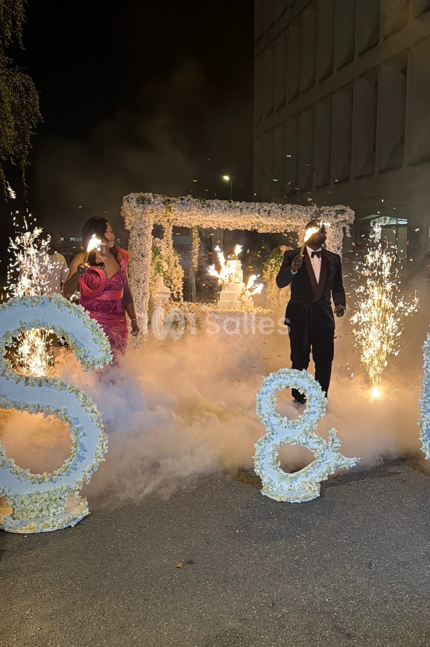 Un couple marche entre des décorations florales et des étincelles, entouré de fumée lors d'une célébration nocturne.