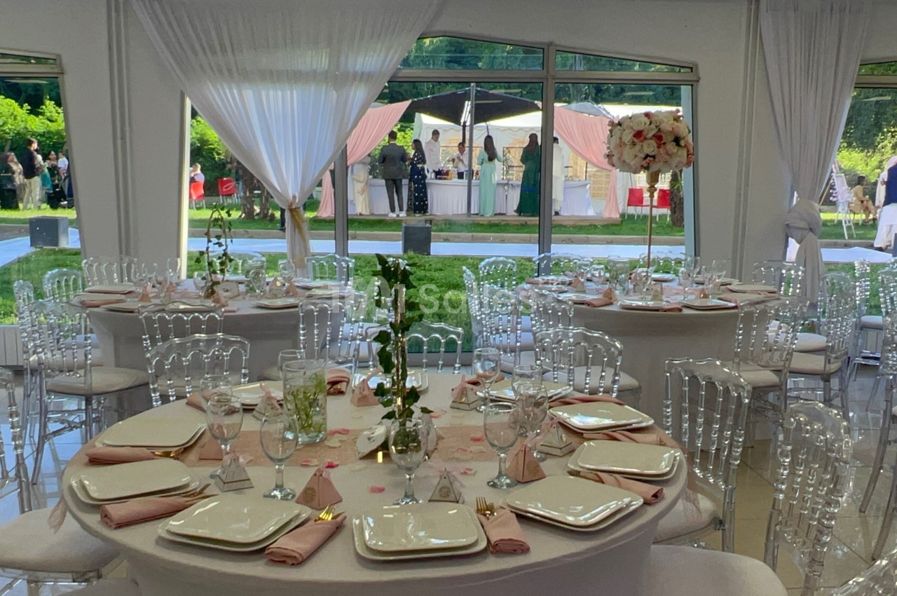 Salle de réception décorée avec des tables rondes, nappes blanches, chaises transparentes et vue sur un jardin extérieur.