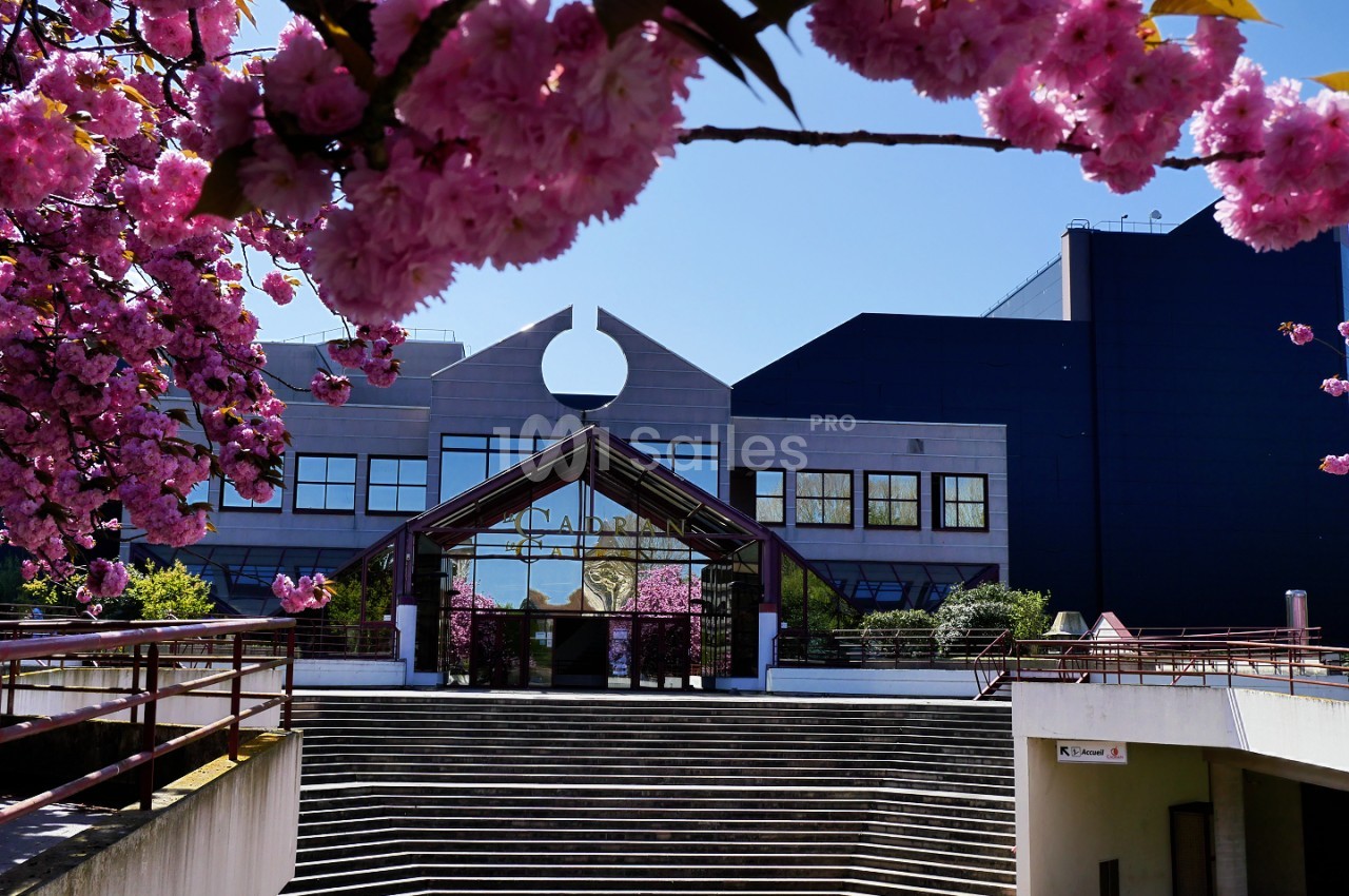 Bâtiment moderne aux façades blanches et noires avec entrée vitrée, entouré de fleurs de cerisier rose en fleurs.