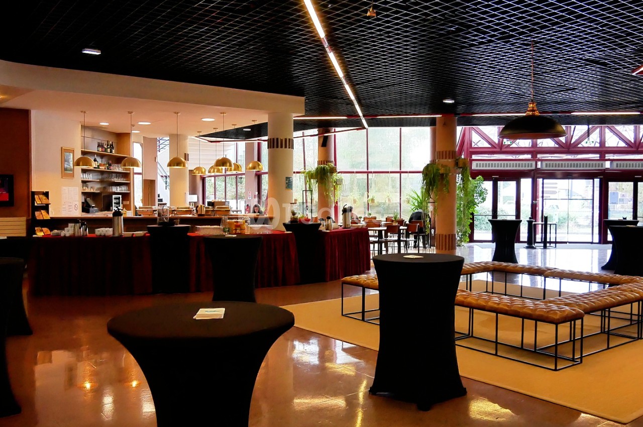 Salle de réception avec sol poli, tables bistrot noires, bar et éclairage moderne - larges baies vitrées.