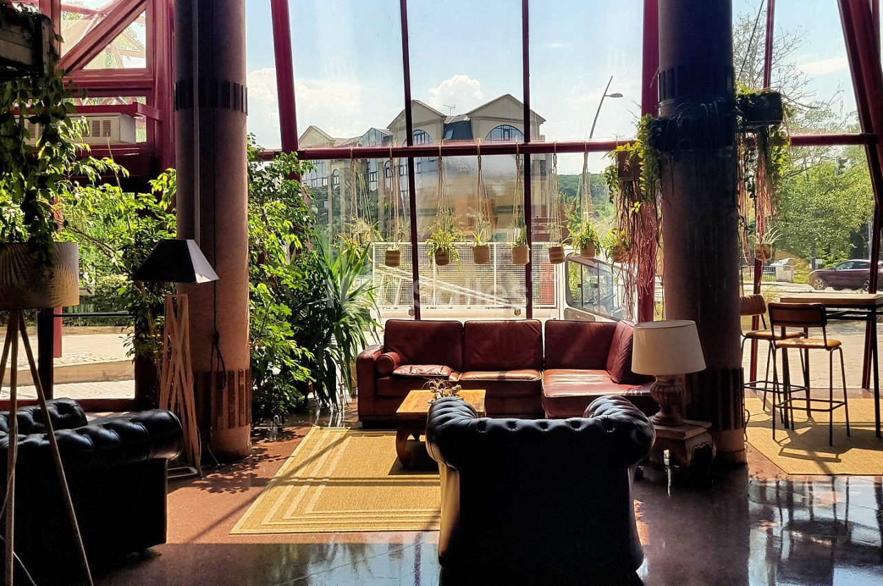 Salle avec canapés bordeaux et noirs, colonnes métalliques, grandes baies vitrées donnant sur paysage boisé.