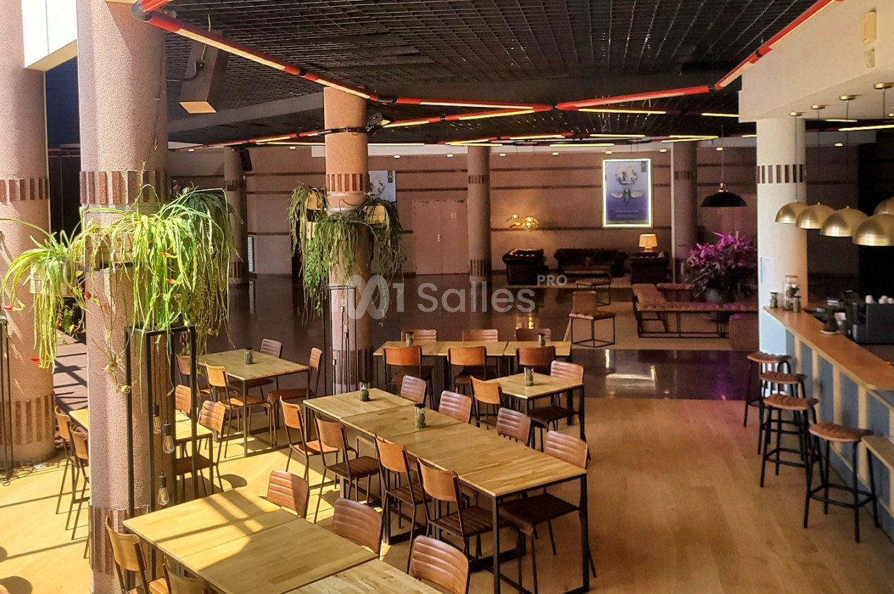 Salle de réception avec tables hautes en bois, chaises roses, plantes vertes, éclairage néon rouge au plafond et bar en arrière-plan.