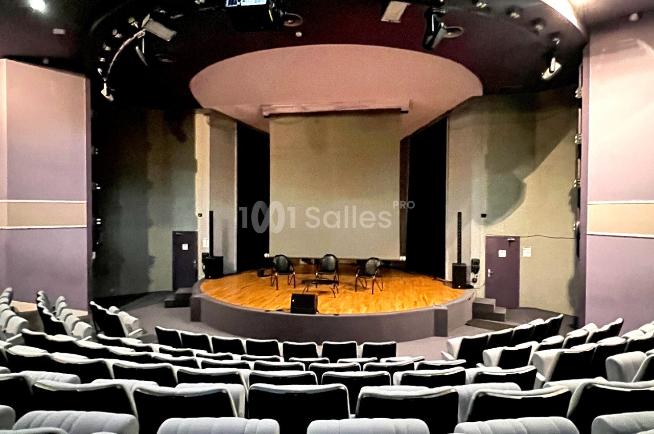 Salle de conférence avec scène circulaire en parquet, chaises noires face à la scène et murs gris-beige avec écran de projection.