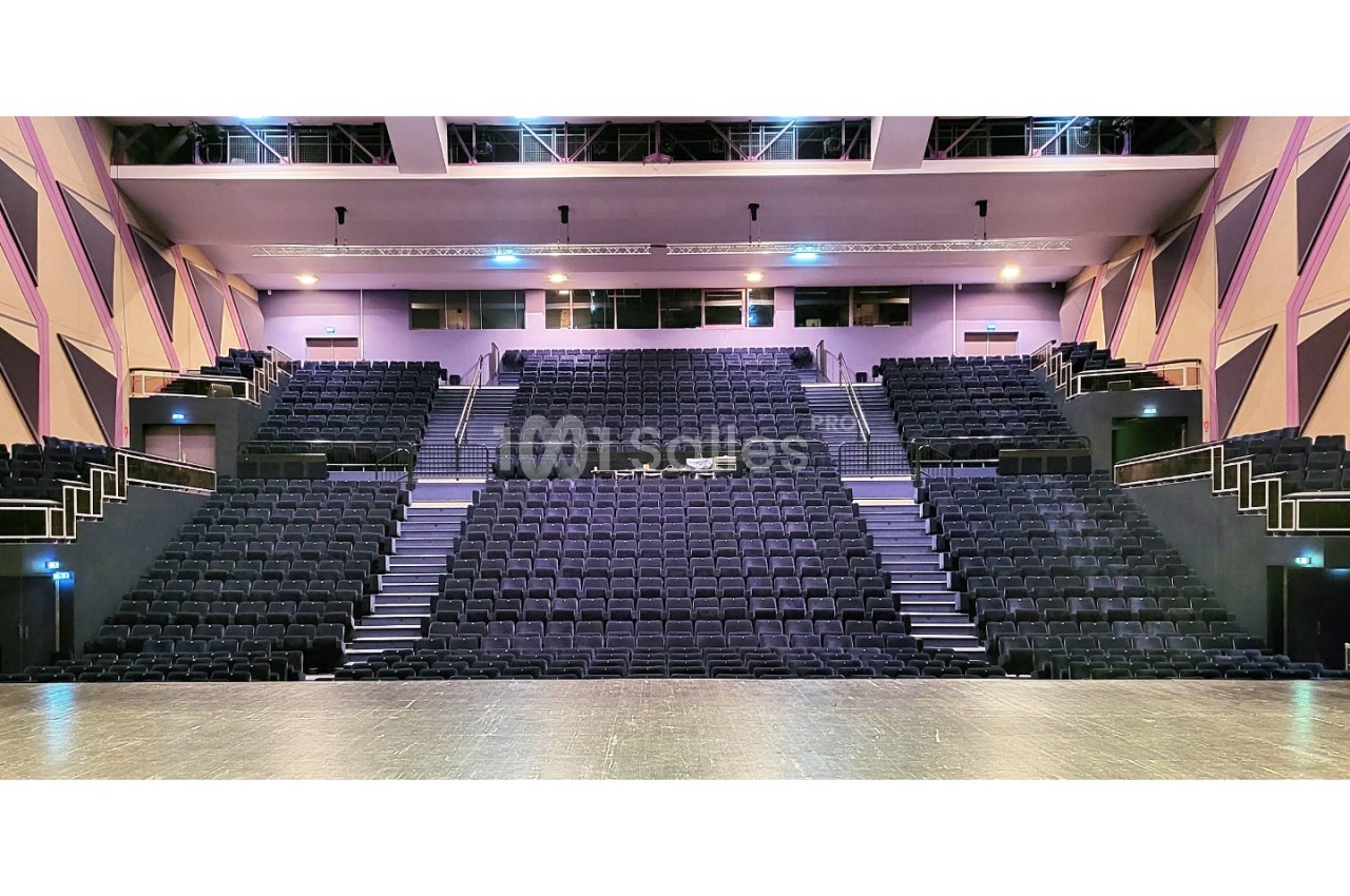 Auditorium avec gradins en escalier, fauteuils noirs, éclairage rose-bleu, sol en parquet clair, capacité plusieurs centaines de places.
