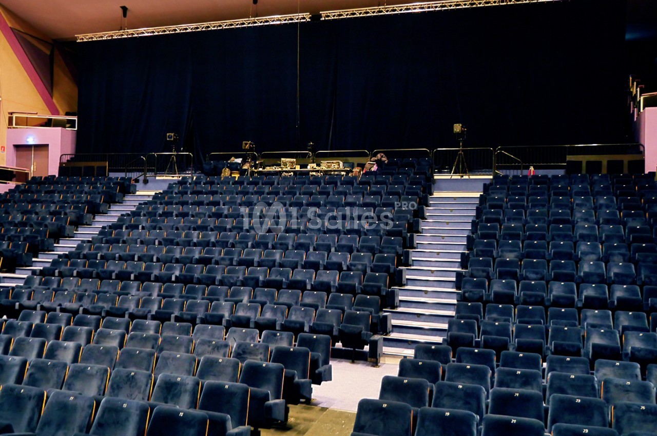 Salle d'auditorium avec fauteuils bleu marine numérotés en gradins, scène équipée de projecteurs et structures métalliques.
