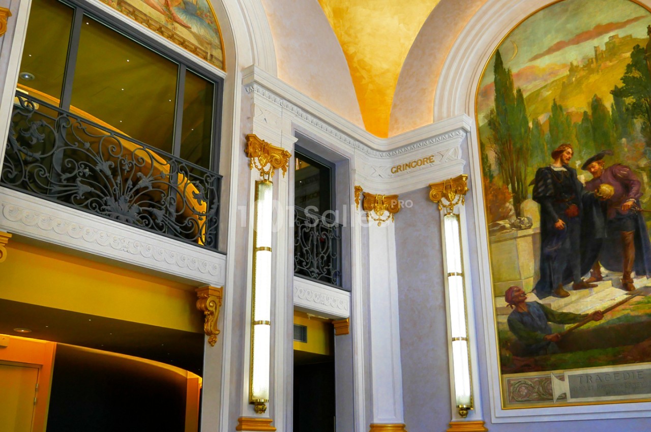 Intérieur aux murs blancs ornés de peintures murales, plafond voûté jaune, appliques dorées et balcon à balustrade noire.