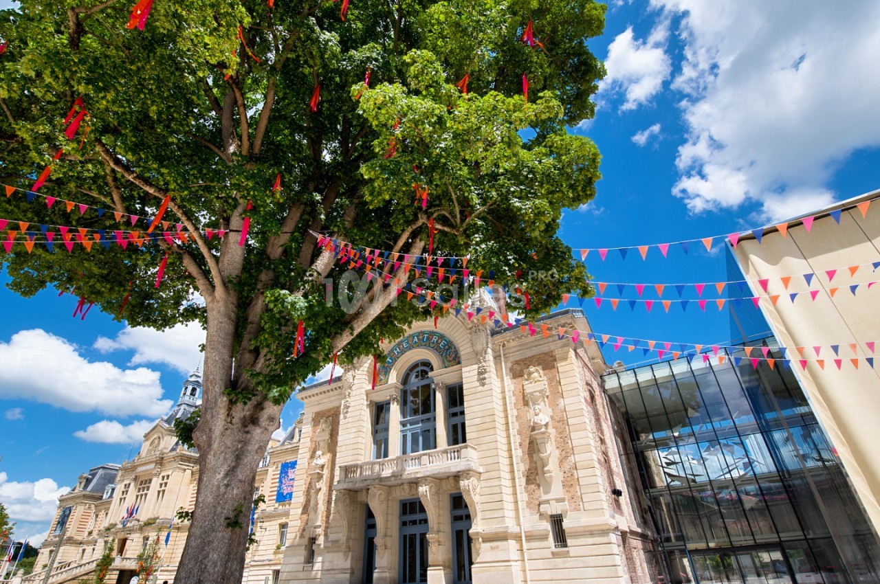 Grand arbre avec rubans rouges devant bâtiment historique en pierre et extension moderne, ciel bleu.