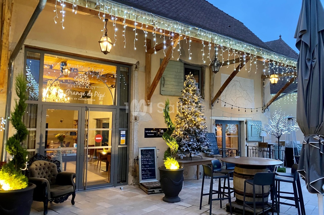 Entrée d'un restaurant décorée pour les fêtes avec guirlandes lumineuses, sapin enneigé et mobilier de terrasse.