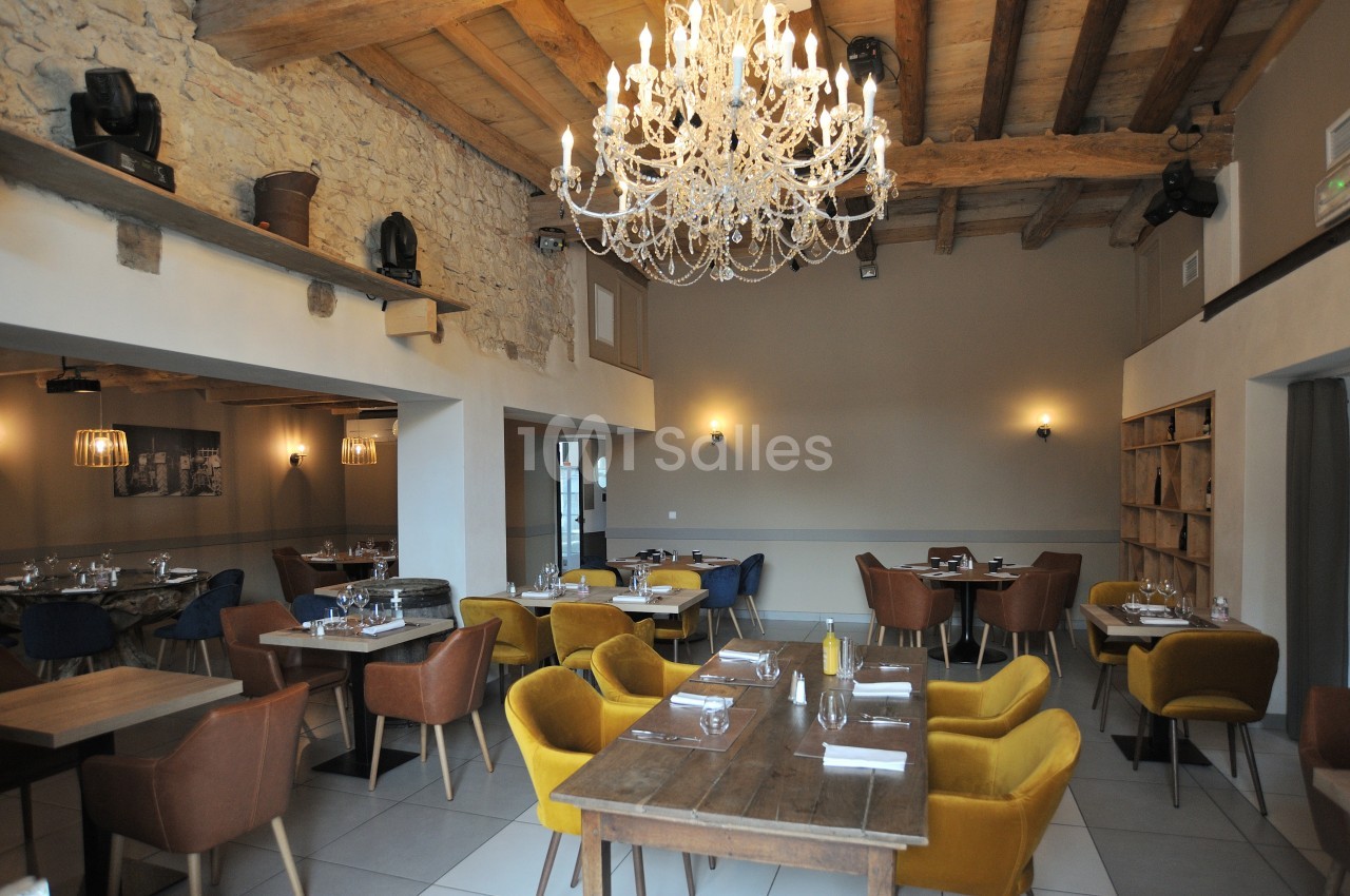 Salle de restaurant lumineuse avec murs en pierre, poutres apparentes, lustre en cristal et mobilier moderne coloré.