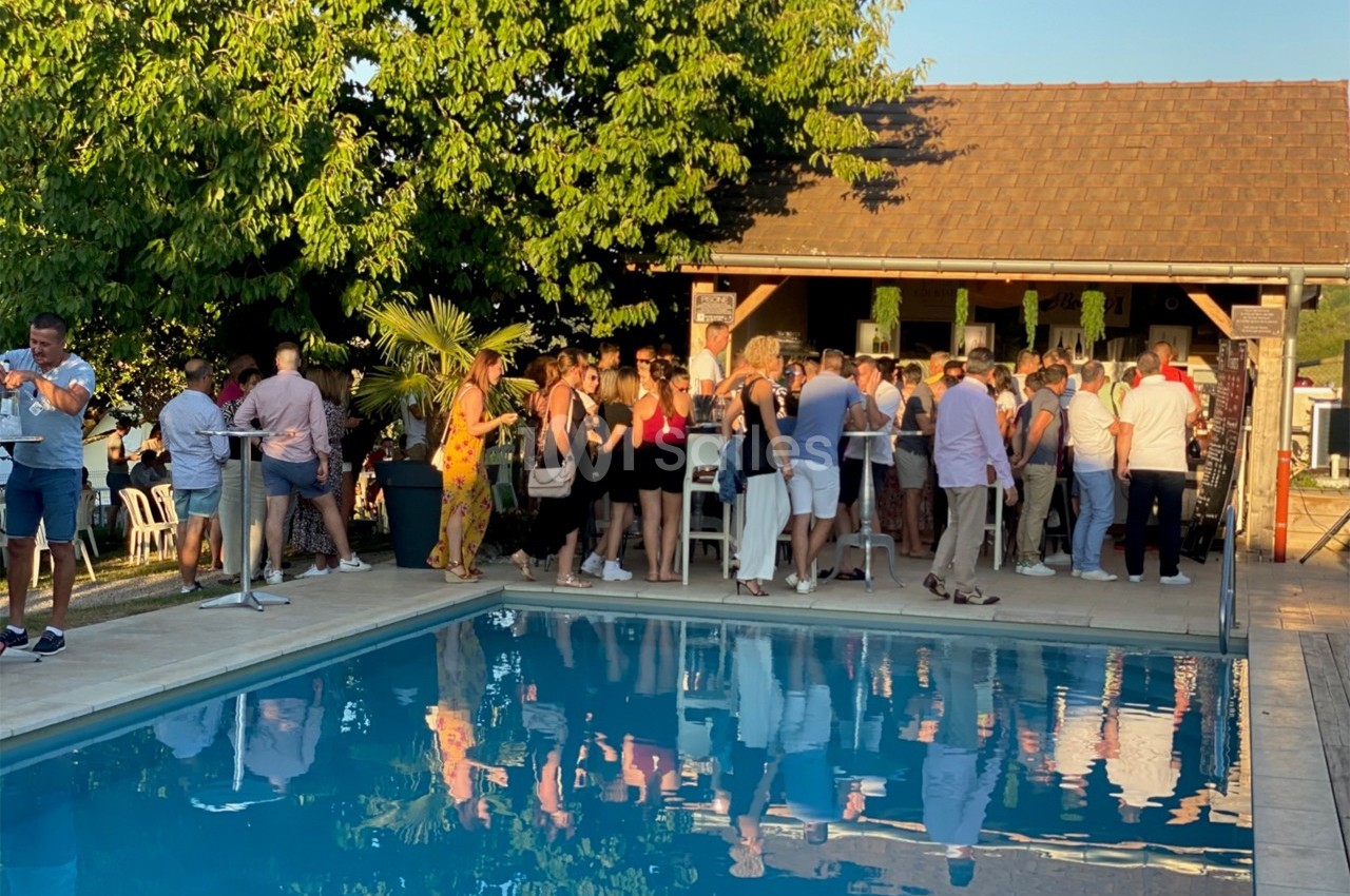 Groupe de personnes rassemblées près d'une piscine, devant un bar extérieur, sous un ciel dégagé en fin de journée.
