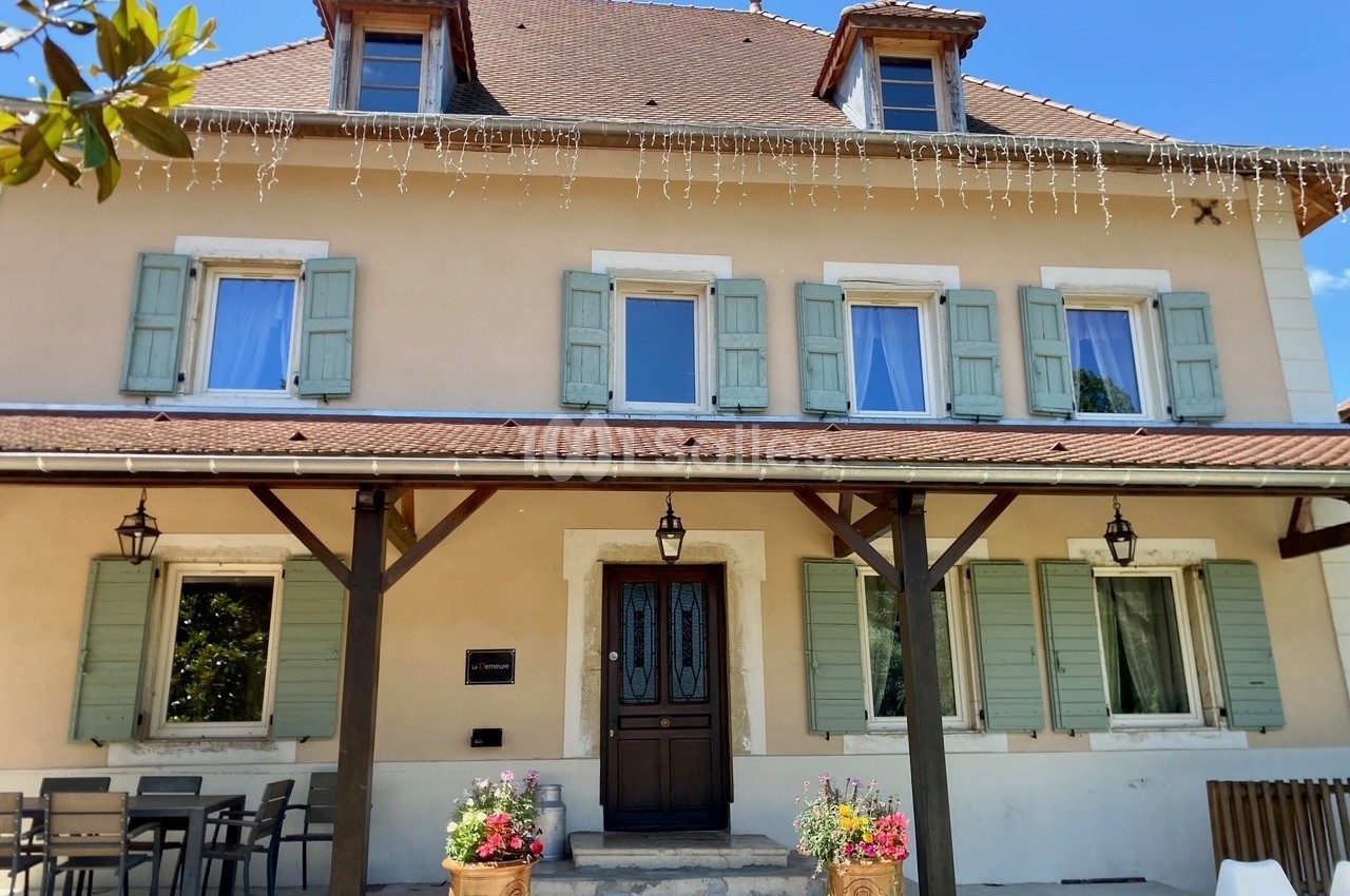 Façade d'une maison beige à volets verts, avec une porte centrale, des fleurs en pot et une table en extérieur.