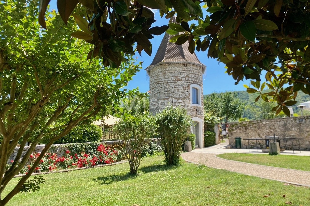 Tour en pierre entourée de verdure, avec des arbres, des fleurs et un chemin dans un jardin ensoleillé.