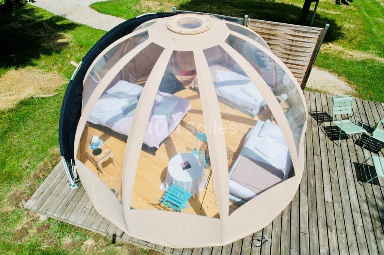 Vue aérienne d'une bulle transparente aménagée avec deux lits, une table et des chaises sur une terrasse en bois.