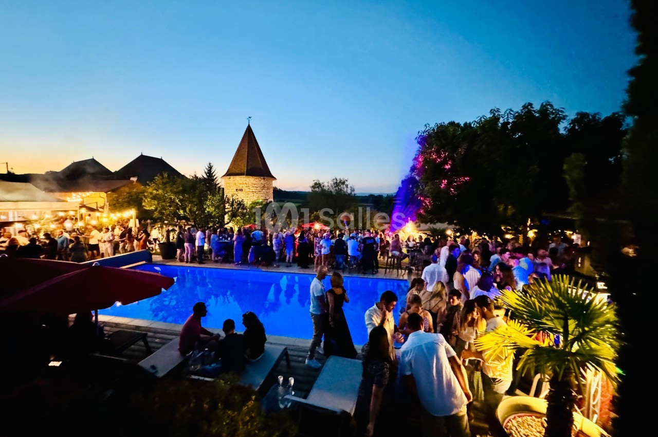 Foule rassemblée autour d'une piscine éclairée en soirée, avec un feu d'artifice en arrière-plan et un bâtiment en pierre.