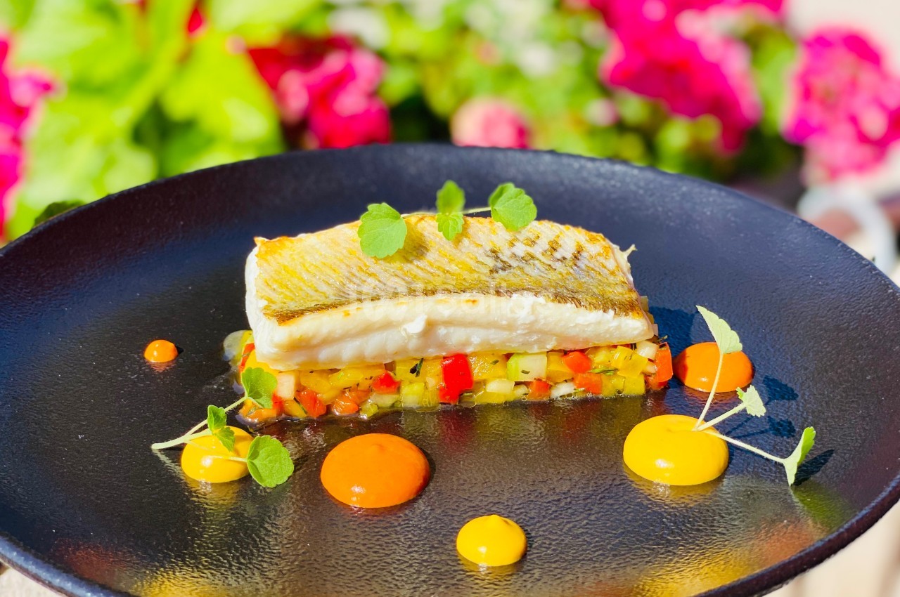 Filet de poisson grillé sur lit de légumes colorés, décoré de sauces et herbes, avec un fond de fleurs floues.