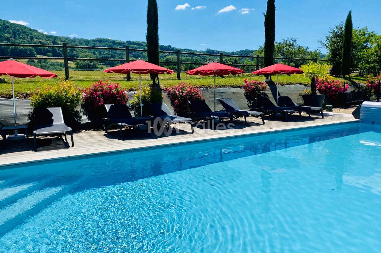 Piscine extérieure entourée de chaises longues, parasols rouges et végétation, avec vue sur des collines verdoyantes.