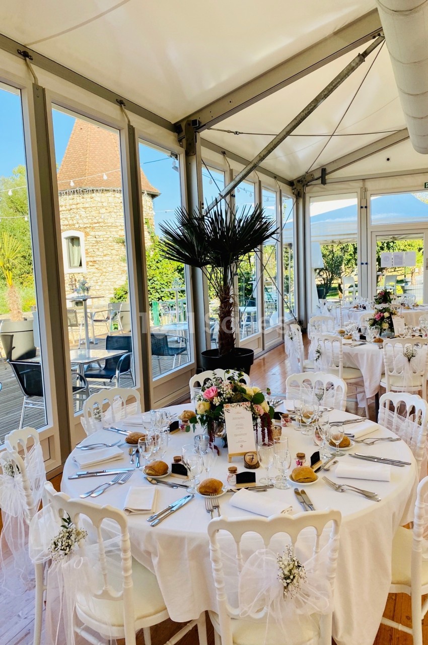 Salle de réception lumineuse avec tables rondes décorées pour un événement, vue sur une terrasse et un jardin.