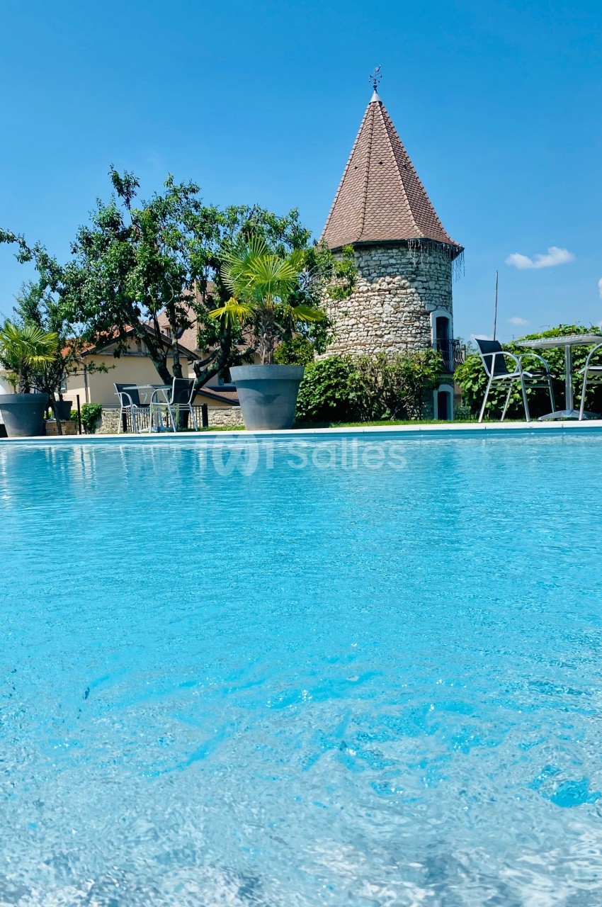 Piscine extérieure avec eau turquoise, entourée de chaises, plantes en pots et un bâtiment en pierre avec toit en pointe.