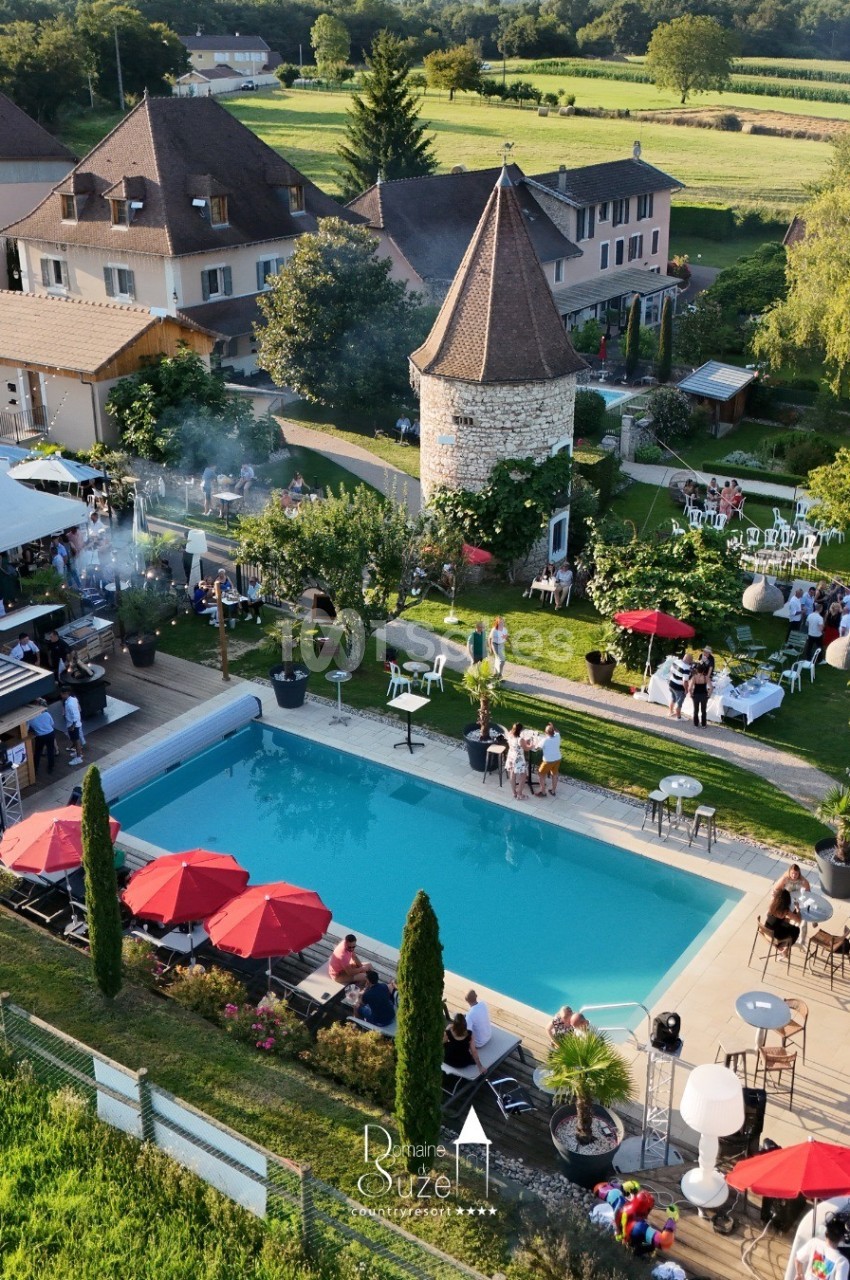 Vue aérienne d'une propriété avec piscine, terrasse, parasols rouges, et bâtiments entourés de verdure.