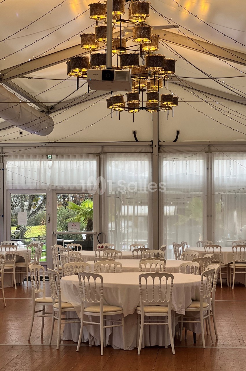 Salle de réception lumineuse avec tables rondes dressées de nappes blanches, sous un plafond décoré de guirlandes lumineuses.
