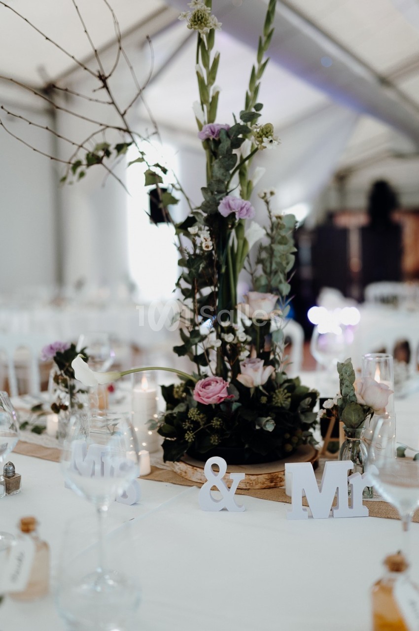 Centre de table floral avec roses et feuillages, décoré de lettres ’Mr & Mrs’ dans une salle de réception lumineuse.