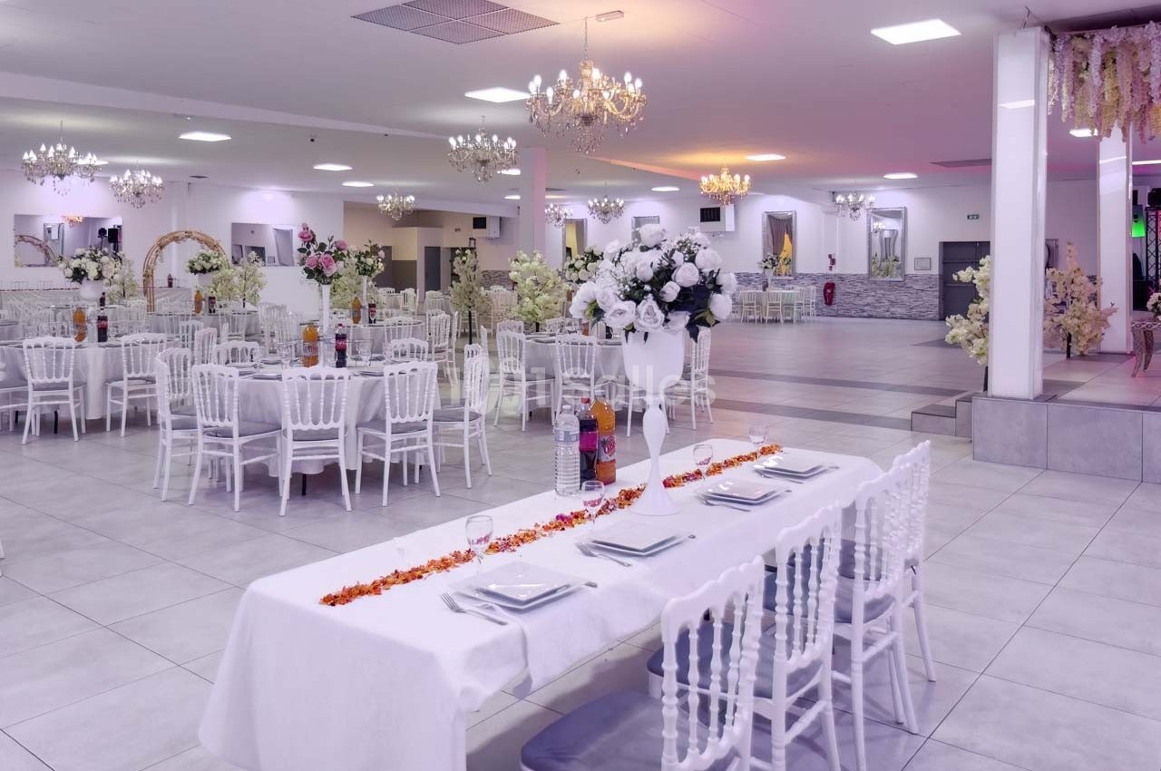 Salle de réception élégante avec tables dressées, chaises blanches et décorations florales sous des lustres lumineux.