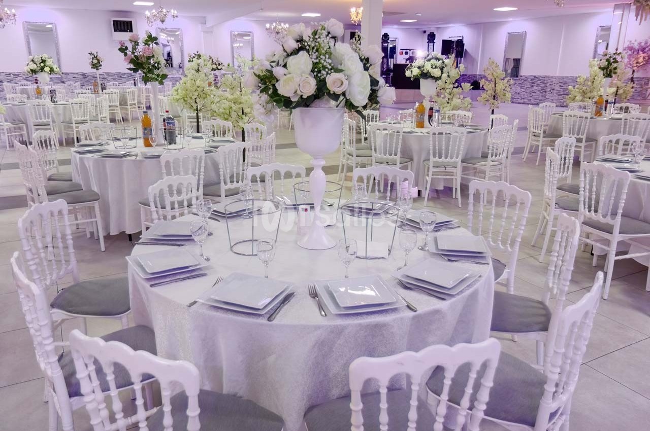 Salle de réception décorée avec des tables rondes, nappes blanches, chaises élégantes et centres de table fleuris.