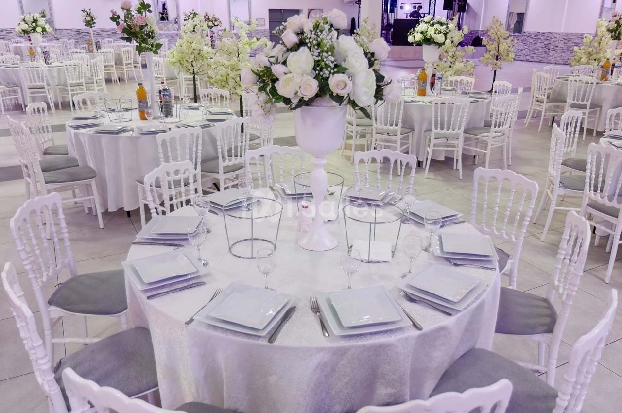 Salle de réception avec tables rondes décorées de nappes blanches, centres de table fleuris et chaises blanches.