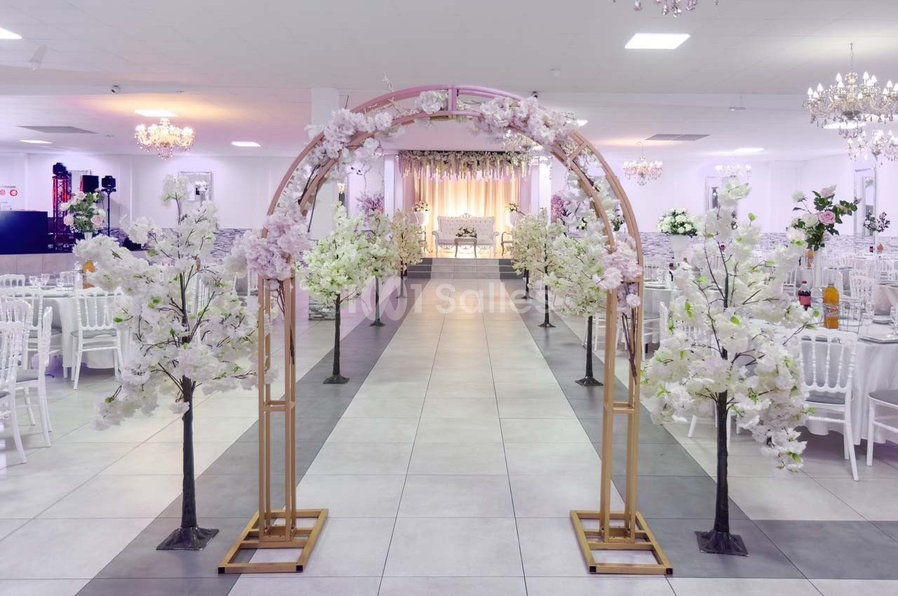 Arche décorée de fleurs blanches dans une salle de réception lumineuse avec des chaises et des tables alignées.