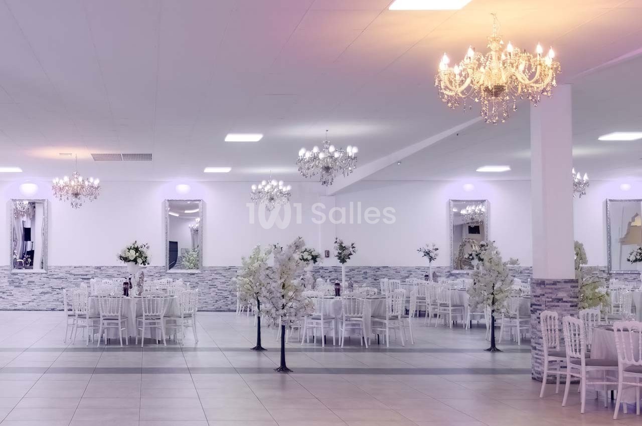 Salle de réception lumineuse avec tables décorées, chaises blanches et lustres élégants suspendus au plafond.