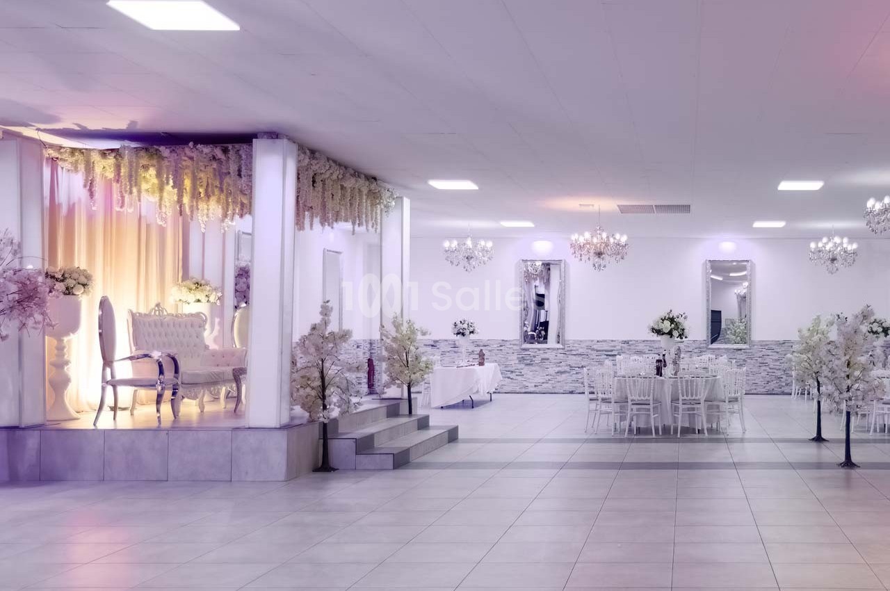 Salle de réception lumineuse avec décorations florales, chaises blanches et espace pour événements.