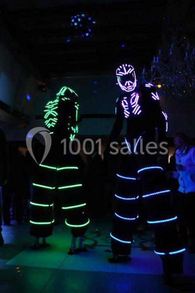 Deux personnes sur échasses portent des costumes lumineux avec des motifs fluorescents dans une ambiance sombre.