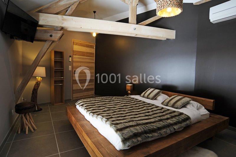Chambre avec lit double en bois, couverture à motif, poutres apparentes, mur sombre et éclairage tamisé.