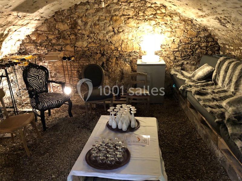 Salle voûtée en pierre avec mobilier éclectique, table dressée de verres et carafes, éclairée par une lampe.