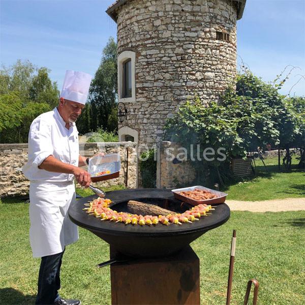 Un chef en tenue blanche cuisine des brochettes et de la viande sur un barbecue rond dans un jardin près d'une tour en…