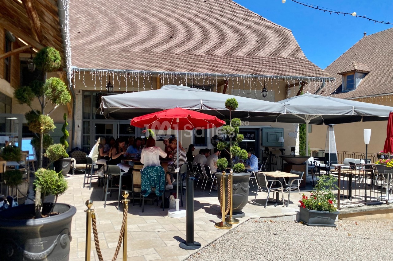 Terrasse extérieure d'un restaurant avec des tables occupées, parasols gris et rouge, et décorations végétales.