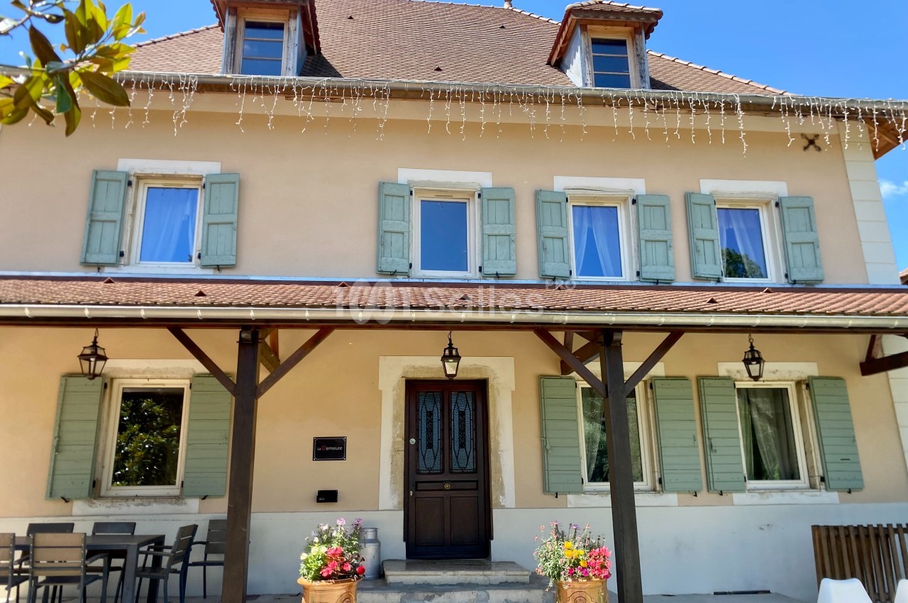 Façade d'une maison beige à volets verts, avec un toit en tuiles, des fleurs en pots et une table sur la terrasse.
