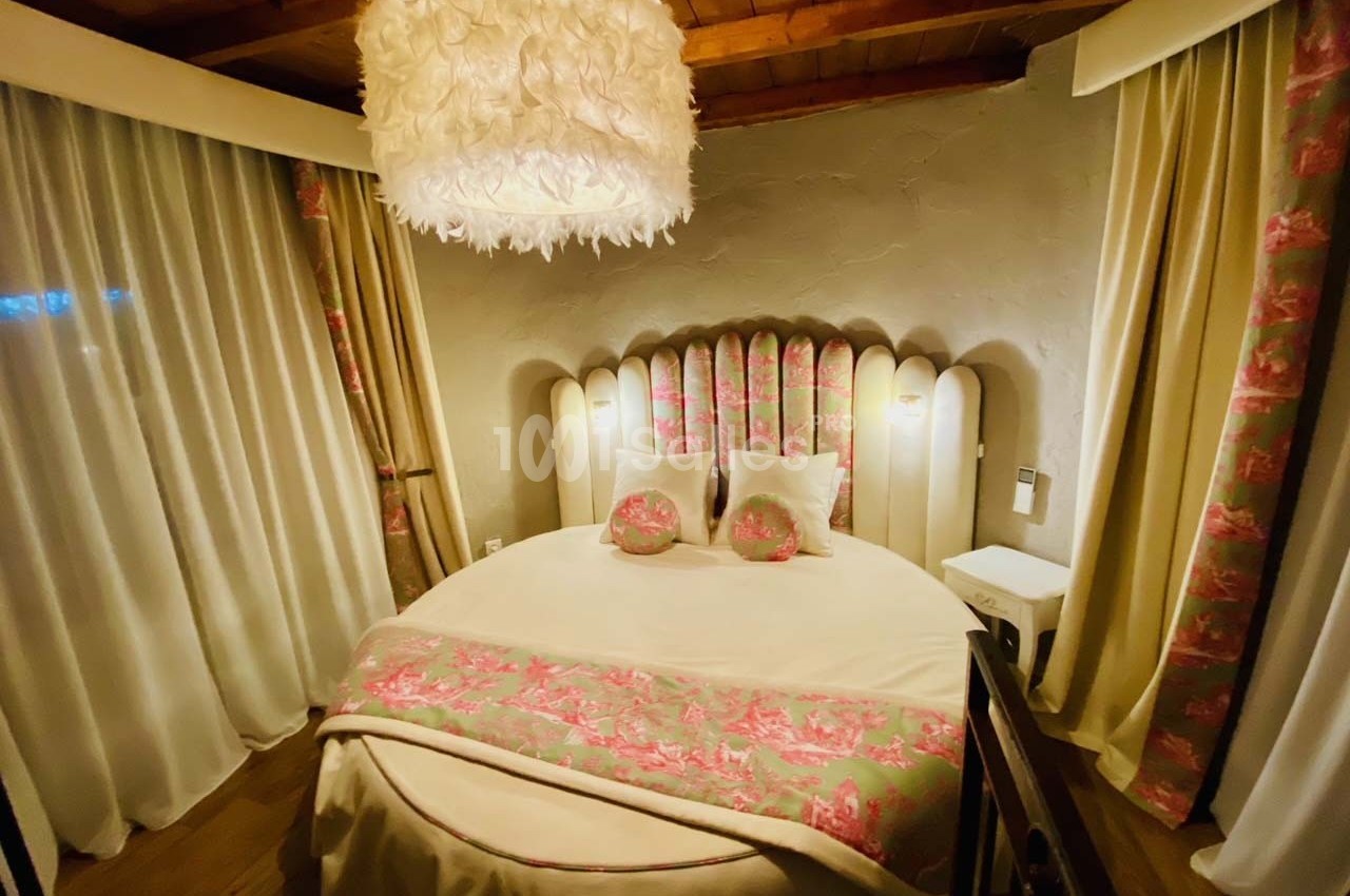 Chambre avec lit rond, tête de lit rembourrée, rideaux fleuris et suspension lumineuse en plumes.