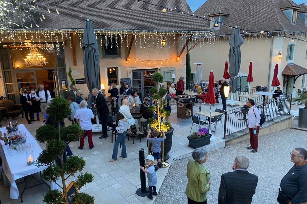 Groupe de personnes rassemblées dans une cour extérieure éclairée, avec tables, parasols et décorations lumineuses.