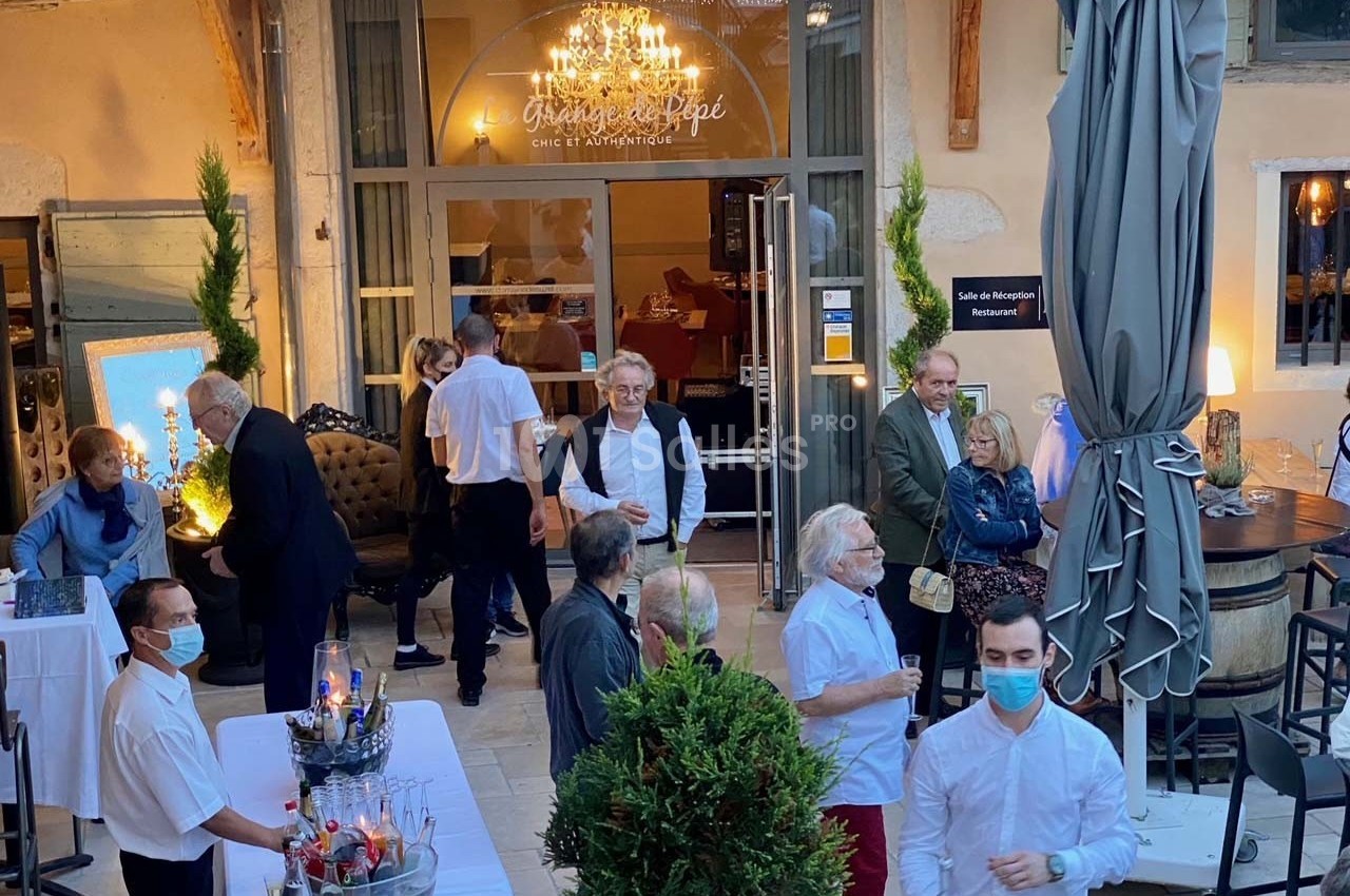Groupe de personnes discutant dans une cour extérieure aménagée avec tables, plantes et éclairage décoratif.