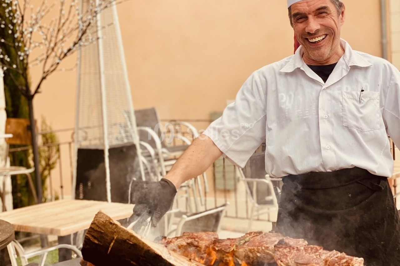 Un cuisinier souriant en tenue blanche prépare des grillades sur un barbecue en extérieur.
