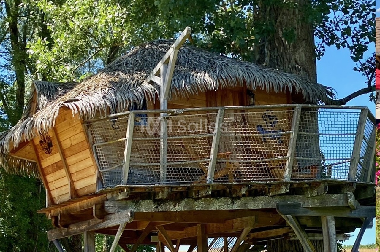 Cabane en bois sur pilotis avec toit en chaume, entourée de filets, située dans un environnement arboré.