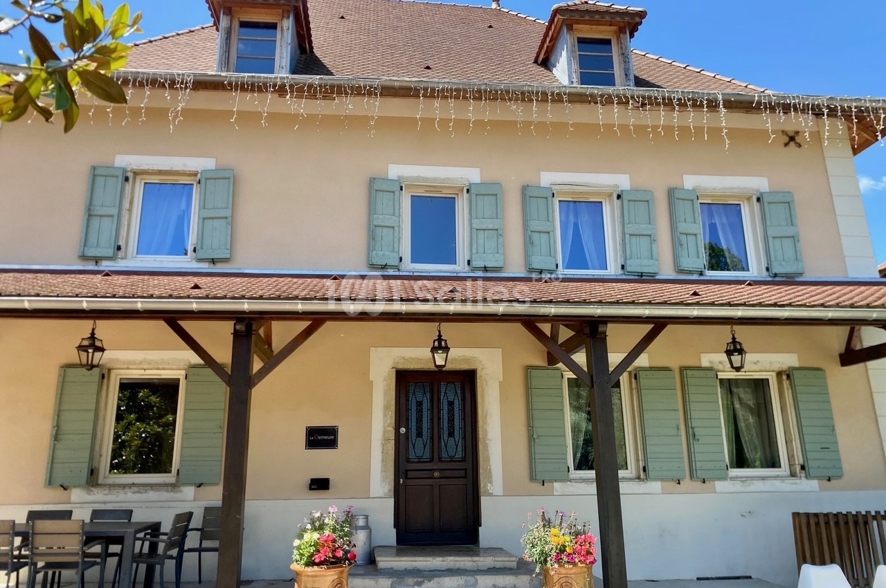 Façade d'une maison beige à volets verts, avec une porte centrale, des fleurs en pot et une table en extérieur.