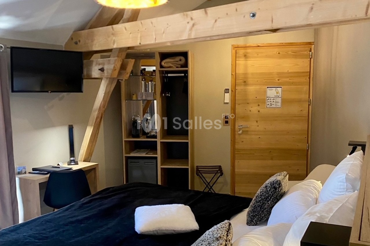 Chambre avec poutres apparentes, lit double, bureau, télévision murale et espace de rangement en bois clair.